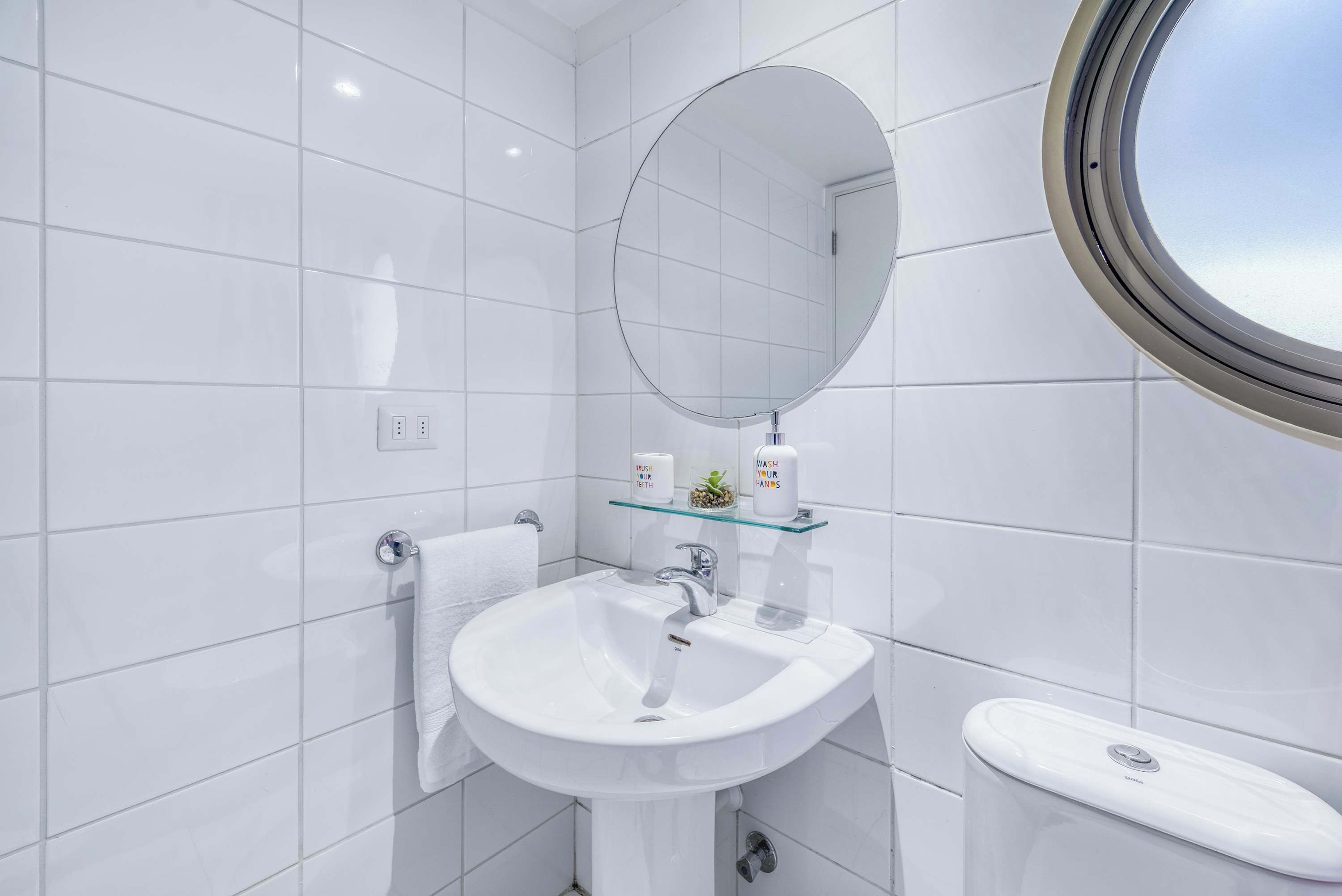 A bathroom with white tiles, a round mirror, a glass shelf with toiletries, and a toilet.