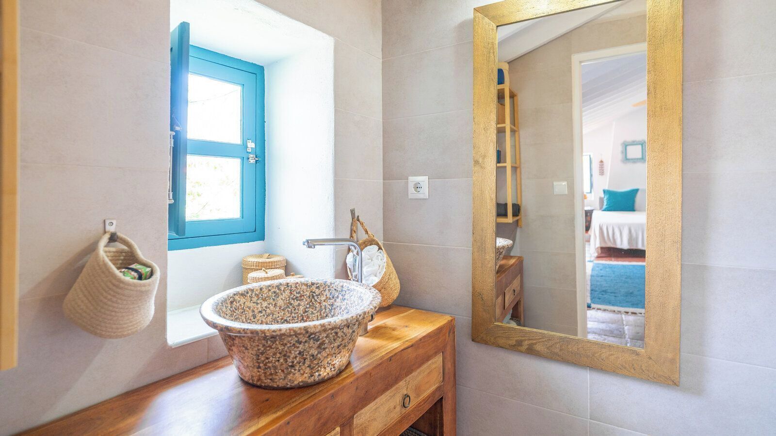 Ein Badezimmer mit einem Becken aus Stein, einem Fenster mit blauem Rahmen, einer Holzkommode und einem Spiegel mit goldener Rahmung.