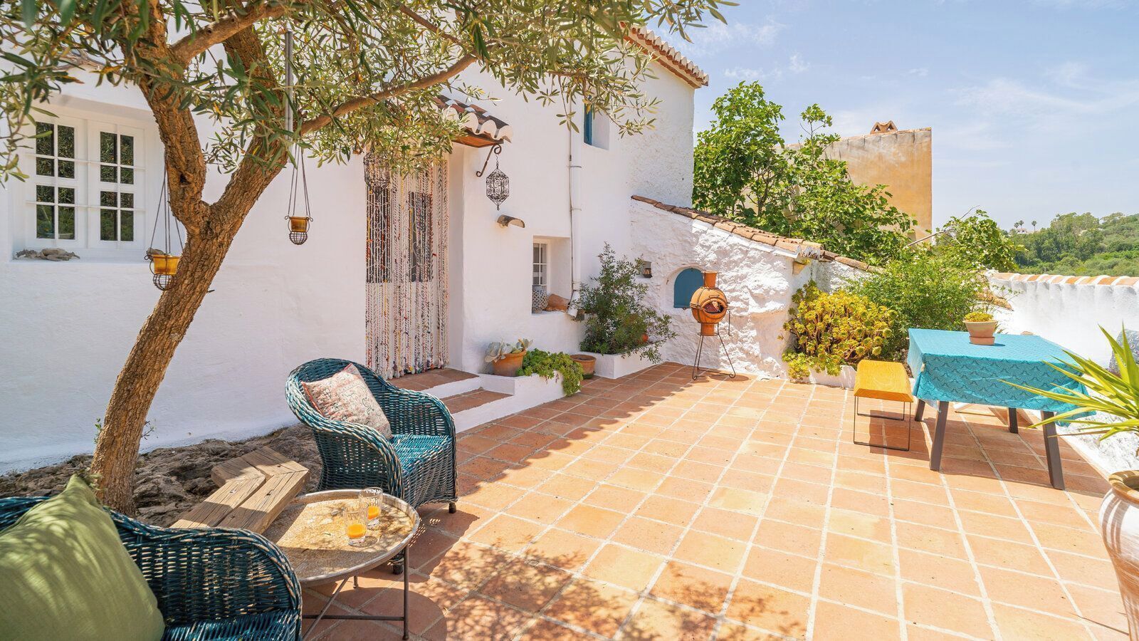 A patio with wicker furniture, a table with drinks, and a white building with a tiled roof.