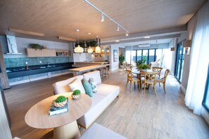 A modern kitchen with a central island, a dining area with wooden chairs, and a large window providing natural light. - Studio Completo - Vila Olímpia (São Paulo)