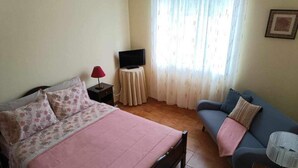 Un dormitorio con cama, sofá, televisión sobre una mesita y una ventana con cortinas.