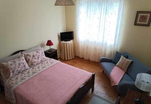 Ein Schlafzimmer mit einem Bett, einem Schlafsofa, einem Fernseher auf einem kleinen Tisch und einem Fenster mit Vorhängen.