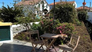 En udendørs terrasse med et bord, stole, en parasol samt en have med blomster og buske.