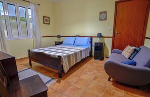 A bedroom with a bed, a sofa, a nightstand, a lamp, a wooden wardrobe, and a window with curtains. - Apartment 2 at Casa da Avo Ines (São Brás)