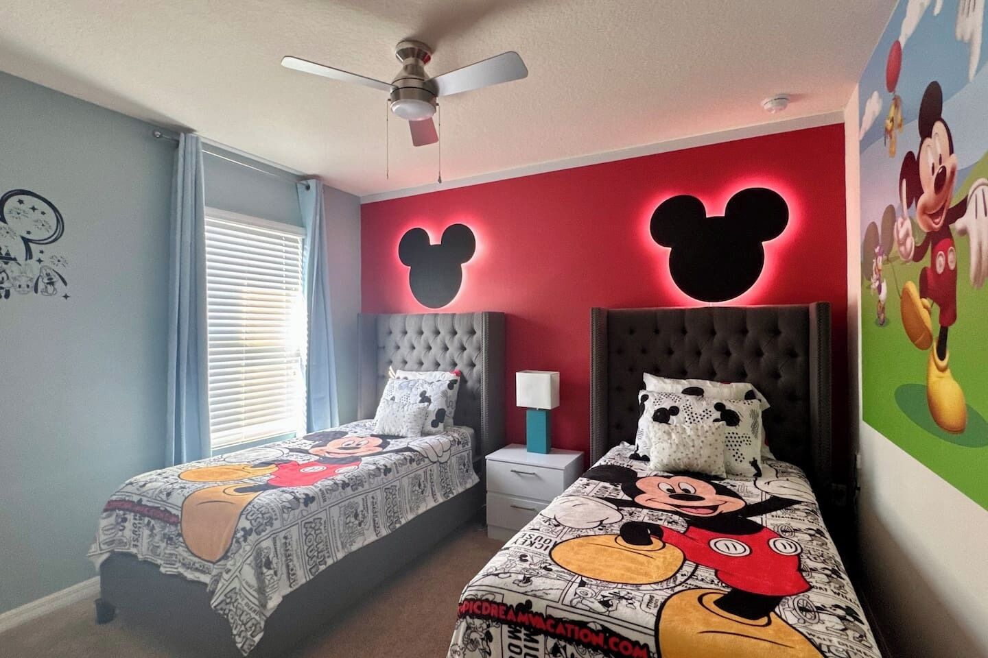 Un dormitorio con dos camas, cada una con ropa de cama temática de Mickey Mouse, un ventilador de techo y una ventana con persianas.