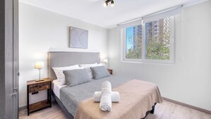 Quarto com cama, mesa de cabeceira, janela e vista para edifícios.