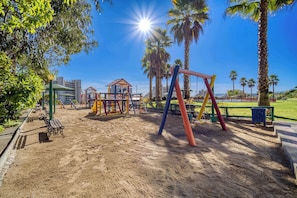 A playground with swings and climbing structures, surrounded by palm trees and a clear sky.