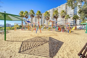 A playground with swings and slides, surrounded by palm trees and a building in the background.