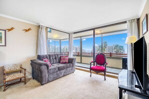 A living room with a grey sofa, a red chair, a wooden bench, a TV, and large windows with a city view.