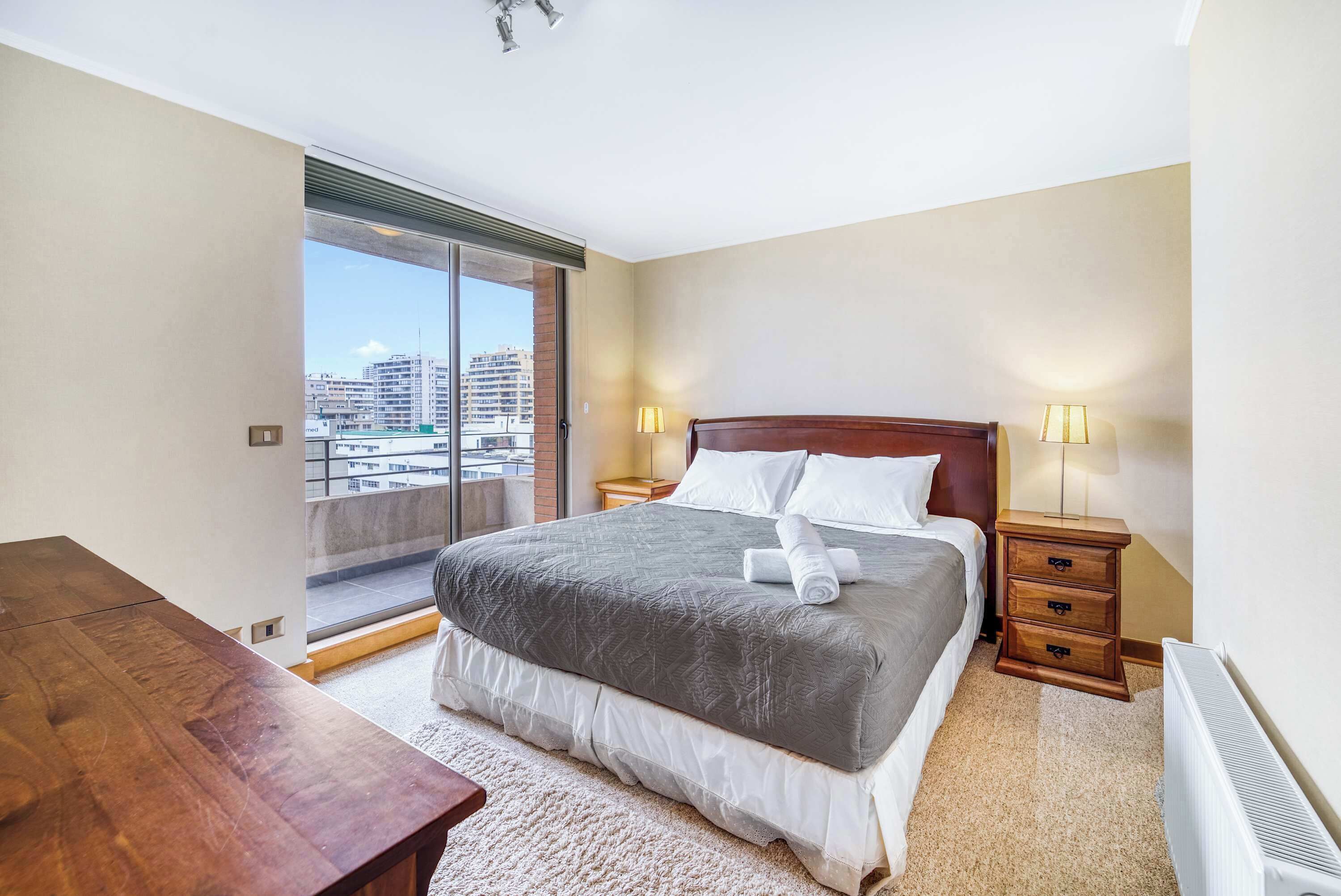 A hotel room with a bed, bedside tables, a desk, and a view of the city through a large window.