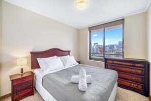 A hotel room with a bed, a nightstand with a lamp, a dresser, and a window with a city view. - Marina Norte apartment 3BR and pool (Viña del Mar)