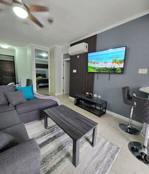 Ein Wohnzimmer mit einem Ecksofa, einem Couchtisch, einem an der Wand montierten Fernseher und einem Deckenventilator.