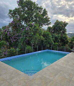 Ein rechteckiger Swimmingpool mit blauen Mosaikfliesen, umgeben von einer Fliesenveranda und einem Zaun.