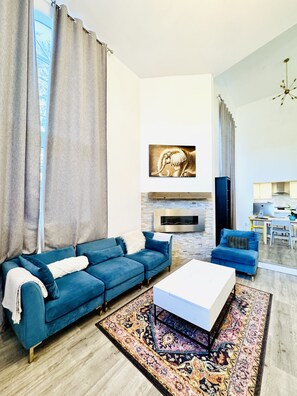 A modern living room with a blue sofa, a white coffee table, a fireplace, and a kitchen area in the background. - Perfect 4-bedroom house with AC & High ceilings for the Summer (Los Angeles)