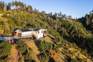 A modern house with a large deck, surrounded by dense forest and hills.