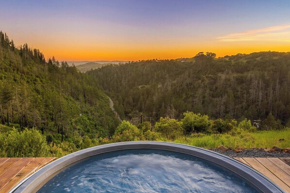 A hot tub with a wooden deck, surrounded by a forested landscape and a view of distant hills.