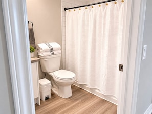 Une salle de bain avec des toilettes, un petit tabouret, un rideau de douche blanc et un plancher en bois.