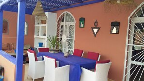A covered outdoor seating area with a table and chairs, a decorative wall, and a large window with bars.