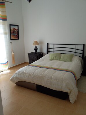 Un dormitorio con cama, mesita de noche, lámpara, un cuadro en la pared y una ventana con cortinas.