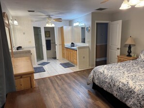 Ein Schlafzimmer mit Bett, Nachttisch, Lampe, Kommode, Spiegel und Deckenventilator, sowie ein Badezimmer mit Dusche, Waschbecken und Toilette.