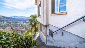 A building with a staircase leading to a balcony, overlooking a valley with mountains in the distance. - Lugano Horizon Retreat (Bigorio)