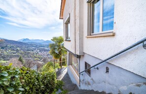 A building with a staircase leading to a balcony, overlooking a valley with mountains in the distance. - Lugano Horizon Retreat (Bigorio)