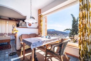 A dining area with a table set for two, wicker chairs, a hanging lamp, and a view of mountains through a large window. - Panorama Stay near Lugano (Bigorio)