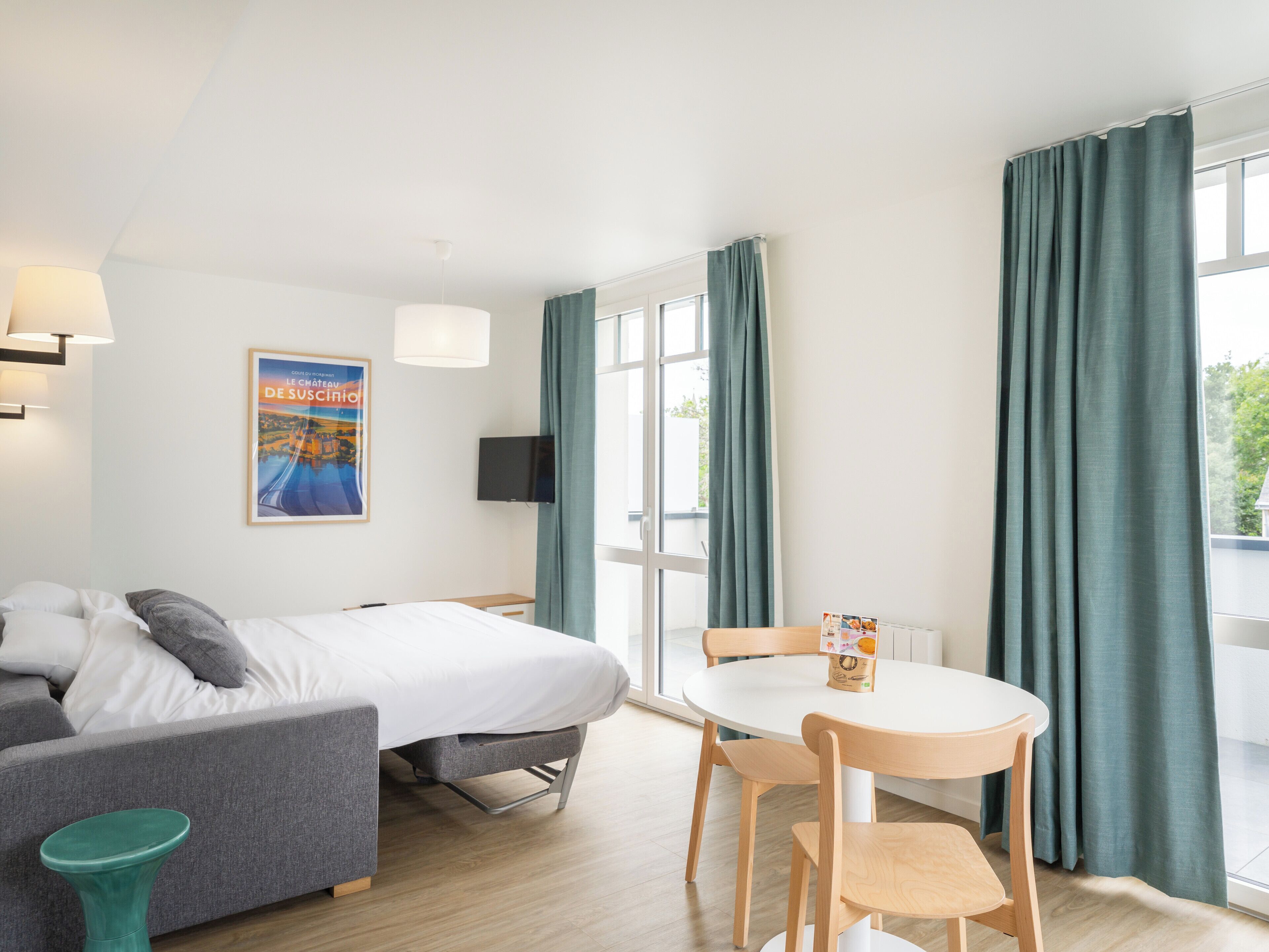 Una habitación de hotel con cama, sofá, mesa de comedor con sillas, televisor, un cartel en la pared y ventanas grandes con cortinas.