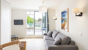 Ein modernes Wohnzimmer mit einer grauen Couch, einem Flachbildfernseher und einem großen Fenster mit Blick auf einen Garten.