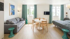 A modern hotel room with a grey sofa, a dining table with chairs, a bed, a TV mounted on the wall, and a window with curtains. - Logis - Grand Privilège - 4p (Sarzeau)