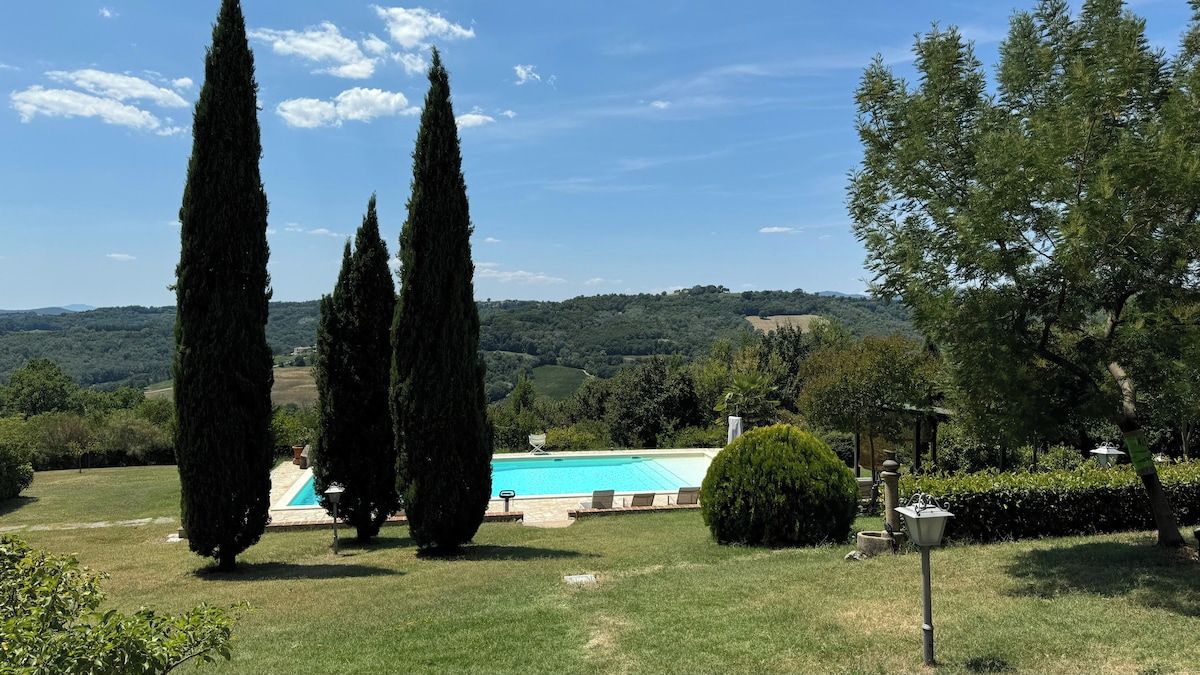 A pool area with tall cypress trees, a well-maintained lawn, and a view of rolling hills in the distance.