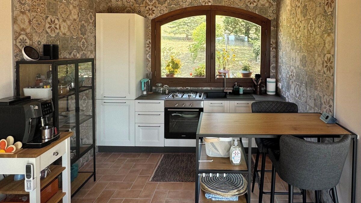 A compact kitchen with a stove, oven, and sink, a dining table with chairs, and a window with a view of greenery.