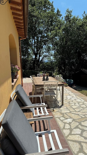 Um pátio externo com mesa e cadeiras, cercado por vegetação e piso de pedra.