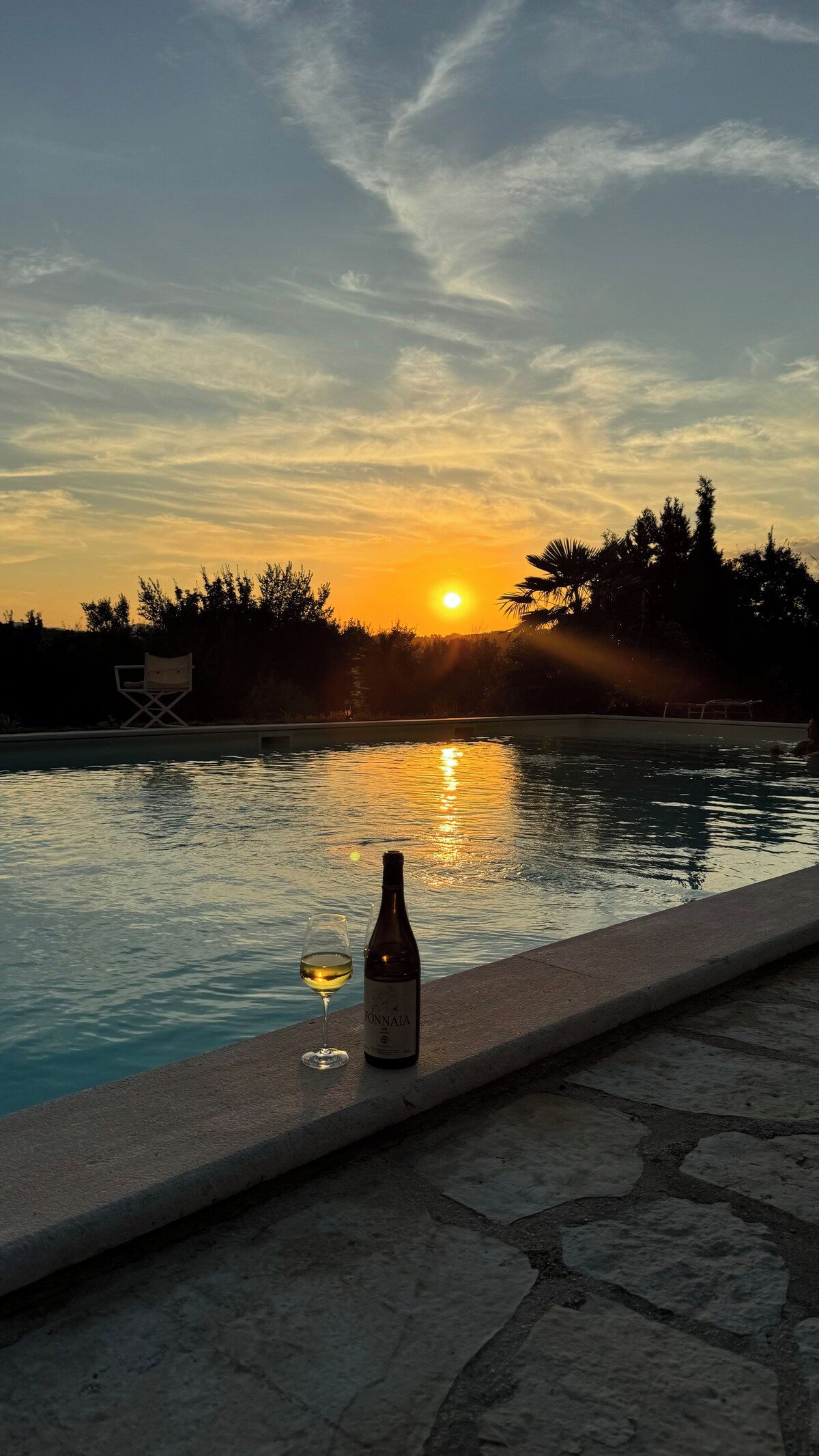 A sunset over a swimming pool with a bottle and glass of wine on the edge.