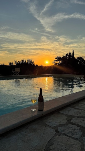 Pôr do sol sobre uma piscina, com uma garrafa e um copo de vinho na borda.