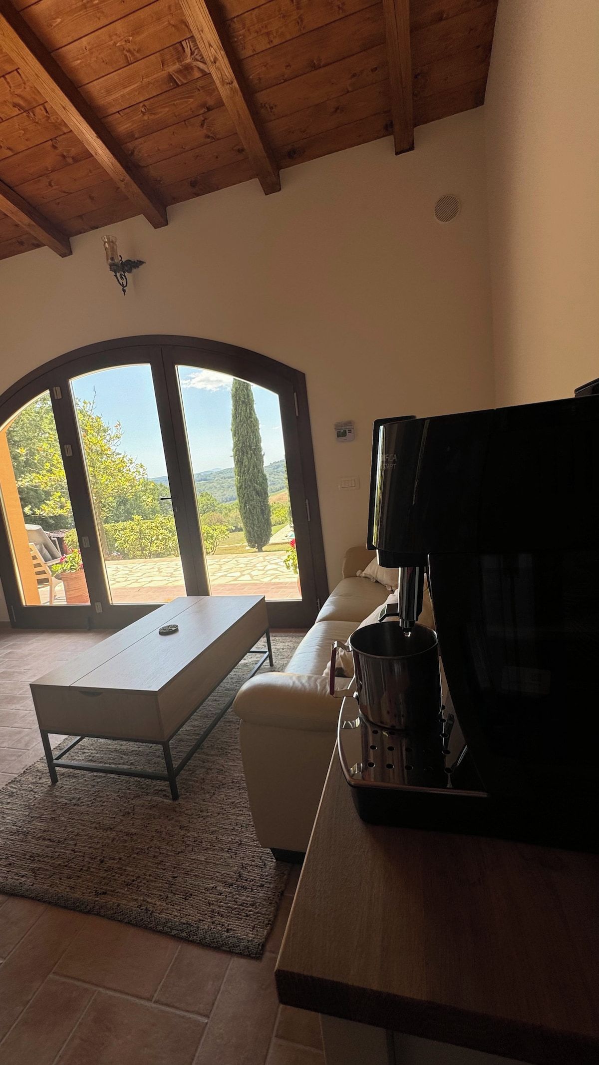 Ein Zimmer mit Holzboden, einer Couch, einem Tisch und einer Kaffeemaschine. Ein großes Fenster bietet Blick auf Grünflächen und einen Baum.