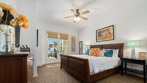 Une chambre avec un lit en bois, un ventilateur de plafond, une table de chevet avec une lampe et une vue sur l’extérieur grâce à une porte vitrée.