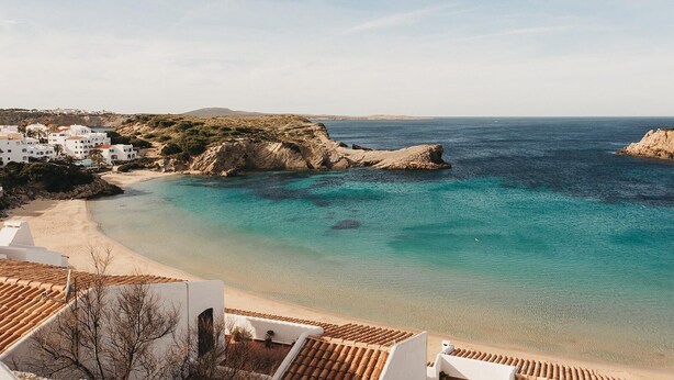 Una vista costiera con spiaggia, acque blu cristalline e edifici bianchi dai tetti in terracotta.