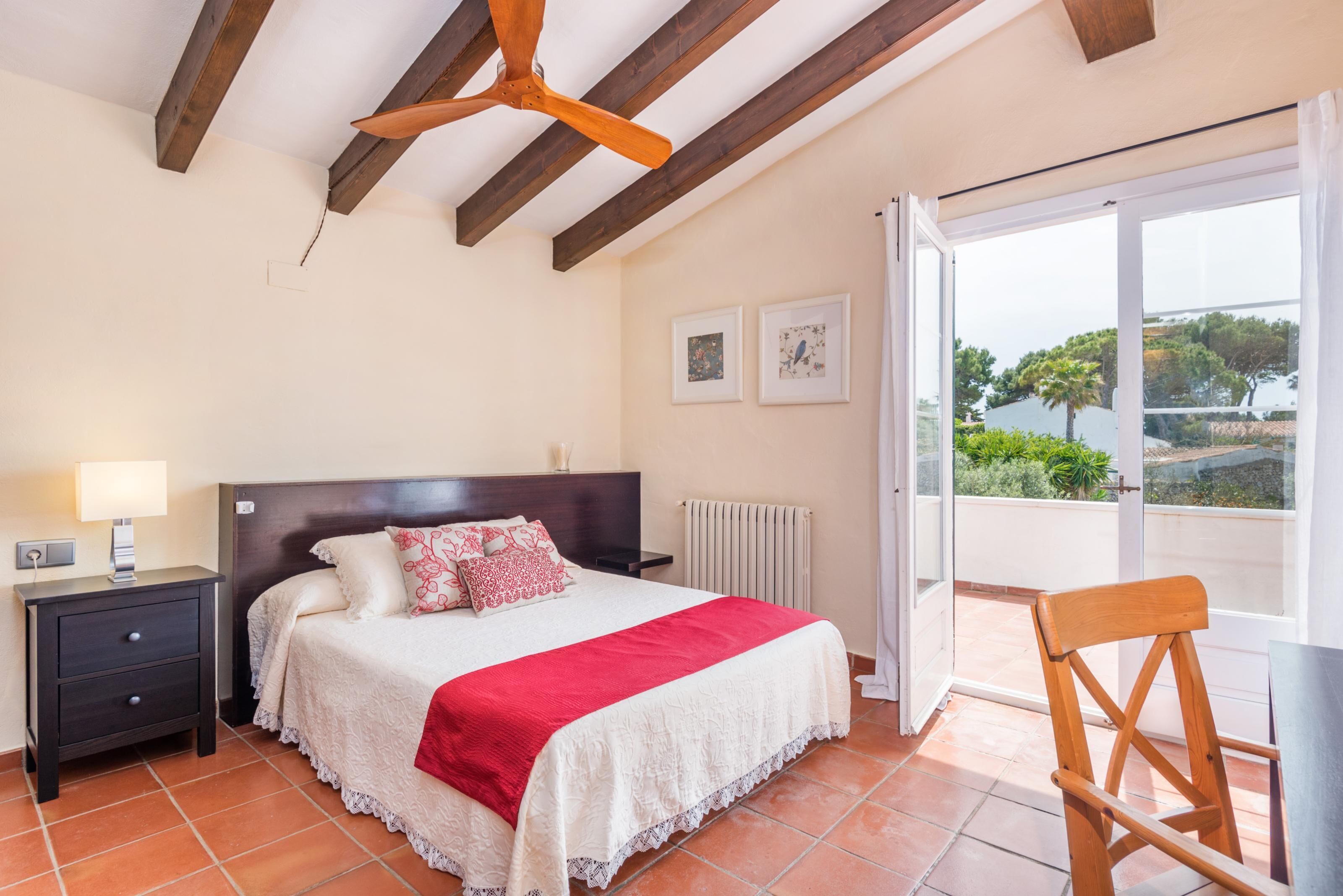 Une chambre avec un lit, une table de chevet, des poutres en bois au plafond, une vue à travers une porte vitrée et une chaise.