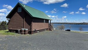 En hytte med grønt tag, grusvej og et vandareal i baggrunden.