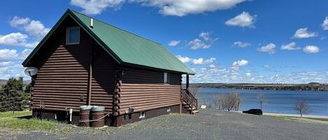 A log cabin with a green roof, a gravel driveway, and a body of water in the background.