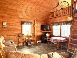 Interior d'una cabana de fusta amb una llar de foc, una televisió, una taula de dinar i un sofà.