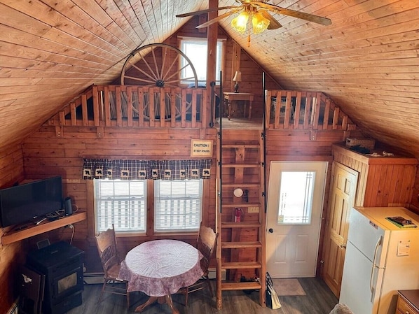 A cozy attic room with a wooden ceiling, a round table with chairs, a television, a refrigerator, and a window with shutters.