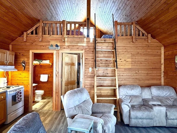 Interior de una cabaña con cocina, comedor, sofá y escalera a un nivel superior.