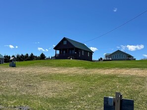 A wooden cabin with a porch, surrounded by a grassy field and a hill.