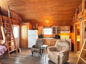 A cozy cabin interior with a wooden interior, a kitchen with a stove and refrigerator, a chair, a small table, and a ladder.