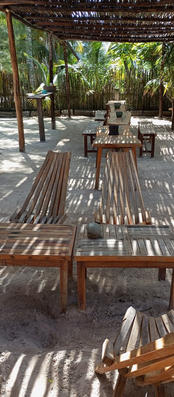Un área de descanso junto a la playa con tumbonas y mesas de madera, rodeada de palmeras y una valla de bambú.