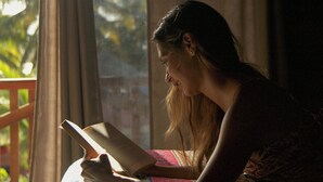 En person læser en bog i sengen med et farverigt sengetæppe, tæt ved et vindue med gardiner.