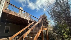 Una escalera de madera que conduce a un balcón con barandilla de metal, rodeado de árboles y un cielo despejado.