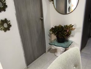 A hallway with a grey door, a round mirror, a small pedestal table with a plant, and a white chair. - Crespito Experience in Cartagena (Cartagena de Indias)